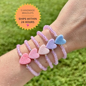 May include: Five beaded bracelets featuring translucent light purple seed beads and a single glittery heart-shaped charm on each. The hearts are colored in shades of pink, light pink, peach, light purple, and light blue, accented with silver beads.