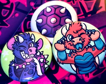 May include: Three colourful button badges featuring cartoon characters. The first badge features a purple and green creature with a spiked collar and a shell on its back. The second badge features a red and blue creature with a spiky shell. The third badge features a red and blue creature with a spiky shell and a big smile.
