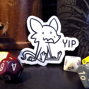 May include: A black and white sticker of a cartoon dog with the word "YIP" next to it. The sticker is on a table with several dice.