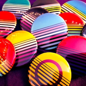 May include: A collection of colorful striped round buttons with black and white stripes. The buttons are arranged in a circle on a purple surface.