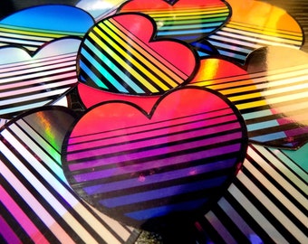 May include: A selection of heart-shaped stickers with a retro design. Each sticker displays a gradient of colours, shifting from red to purple, with black and white stripes. The stickers have a holographic, reflective surface.