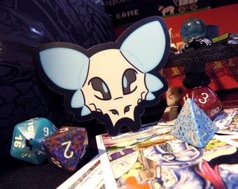 May include: A blue and white sticker of a cartoon skull with large eyes and a toothy grin. The sticker is on a black background with dice and playing cards.