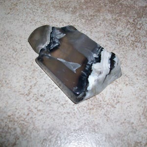 May include: A polished, rectangular stone with a rounded top. The stone features a mix of gray, black, and white colors, with a marbled appearance. The object is likely a decorative piece or a paperweight.