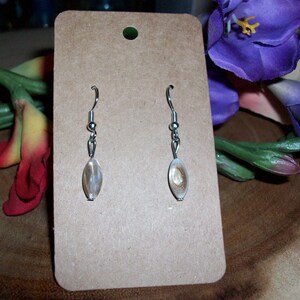 Puede incluir: Un par de pendientes de plata con cuentas ovaladas iridiscentes. Los pendientes se muestran en una tarjeta marrón.