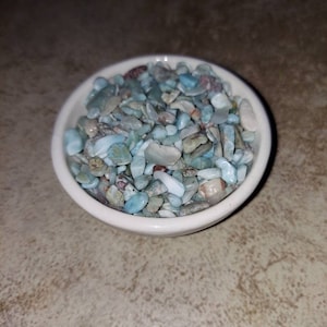 May include: A small white bowl filled with light blue, green, and white tumbled stones. The stones are various sizes and shapes.