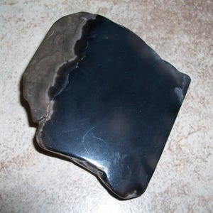 May include: A polished, irregularly shaped, black stone with a lighter gray and brown section on one side. The stone has a smooth, reflective surface and appears to be a natural material. The overall shape is somewhat rectangular with rounded edges.