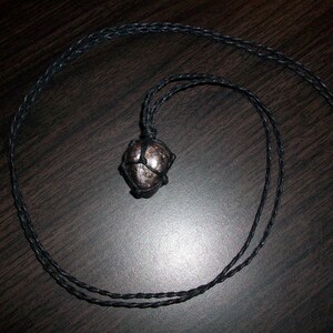 May include: A black cord necklace with a dark grey stone pendant. The stone is wrapped in black cord and has a shiny, reflective surface.