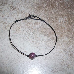 May include: A simple black cord bracelet with a single round, dark purple and green gemstone bead.