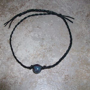 May include: A black braided cord bracelet with a single blue-grey bead at the center. The cord is knotted to secure the bead and has loose ends. The bracelet is simple and minimalist.