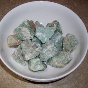 May include: A white bowl filled with green and white rough-cut gemstones. The stones have a mottled appearance and are various sizes.