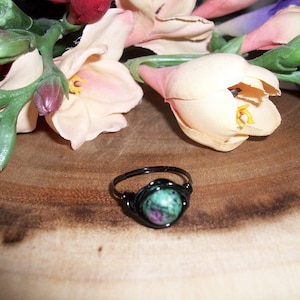 May include: A black wire-wrapped ring with a green and pink gemstone. The ring is sitting on a wooden surface with pink flowers in the background.