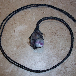 May include: A black cord necklace with a purple and gray stone wrapped in black cord.