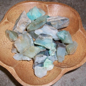 May include: A wooden bowl filled with a variety of green and blue gemstones. The stones are rough and have a natural, unpolished look.