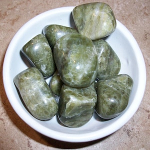 May include: A white bowl filled with several polished green stones. The stones have a smooth, rounded shape and a mottled appearance with lighter green and white inclusions. The bowl is set against a neutral background.
