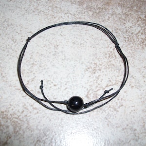 May include: Black beaded bracelet featuring a single, polished, round black bead at the center. The bracelet is made of thin, black cords tied together. The background is a light, speckled surface.