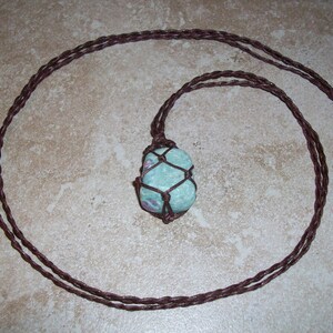 May include: A handmade necklace featuring a light blue stone pendant with red inclusions, encased in a brown macrame net. The necklace has a long, braided brown cord. The stone is approximately 1.5 inches long.