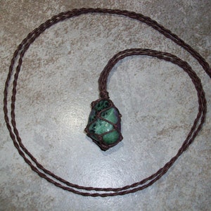 May include: A green and brown gemstone pendant on a brown cord necklace. The gemstone is wrapped in brown cord and has a knotted design.