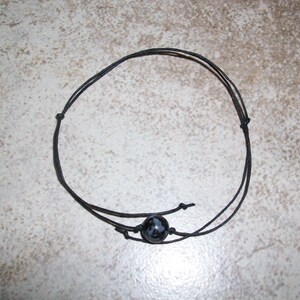 May include: A black cord bracelet featuring a single round, polished black and white snowflake obsidian bead. The bracelet is constructed from thin, dark cord with adjustable sliding knots on either side of the central stone bead.