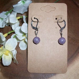 May include: A pair of leverback earrings with a small, round, lavender-colored bead. The earrings are displayed on a brown earring card with a floral arrangement in the background. The earrings are made of silver-colored metal.