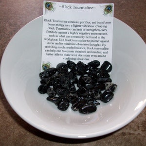 May include: A white bowl filled with black tourmaline stones. A small card with the text "Black Tourmaline" is visible in the background. Black Tourmaline cleanses, purifies, and transforms dense energy into a lighter vibration. Carrying Black Tourmaline can help to strengthen one's fortitude against a highly negative environment, such as what can commonly be found in the workplace. Use black tourmaline to protect against stress and to minimize obsessive thoughts. By providing much needed balance, black tourmaline can help one to remain detached and neutral, and better able to make wise decisions even amidst confusing situations.