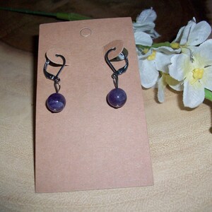 May include: A pair of amethyst gemstone earrings with silver leverback closures. The earrings are displayed on a brown card.