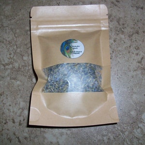 May include: A brown paper bag with a clear window, filled with dried lavender buds. The bag has a circular label with a peacock feather design and the text "Lavender Buds" and "Earth Magick Creations".