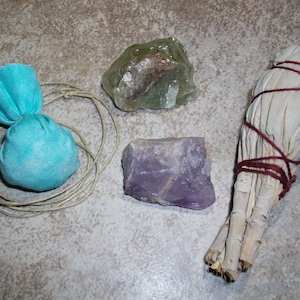 May include: A collection of crystals and herbs for smudging and cleansing rituals. The items include a light blue fabric pouch, a green crystal, a purple amethyst crystal, and a bundle of white sage tied with red string.