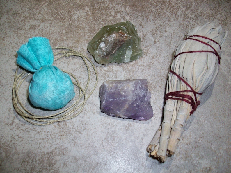 K&ouml;nnte beinhalten: Eine Sammlung von Kristallen und Kr&auml;utern f&uuml;r R&auml;ucher- und Reinigungsrituale. Die Artikel umfassen einen hellblauen Stoffbeutel, einen gr&uuml;nen Kristall, einen violetten Amethystkristall und ein B&uuml;ndel wei&szlig;en Salbeis, das mit roter Schnur gebunden ist.