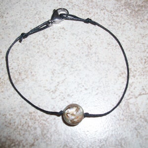 Puede incluir: Un collar de cordón negro con un cierre plateado y una sola cuenta redonda marrón y blanca.