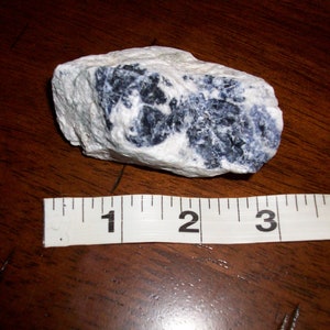May include: A rough, white and blue rock with dark blue veins. The rock is about 2 inches long and is sitting on a wooden surface next to a white measuring tape.