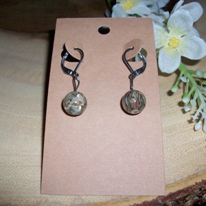 May include: A pair of dangle earrings with a round, textured bead. The beads have a mix of colors, including brown, beige, and hints of blue and green. The earrings are attached to a silver-tone leverback clasp.