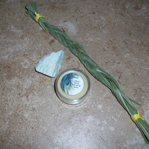 May include: A small jar of yellow balm with a peacock feather design and the text "Wilding Salve" and "Nourishing Balm". A light blue rock and a bundle of dried green grass tied with yellow string are also in the image.
