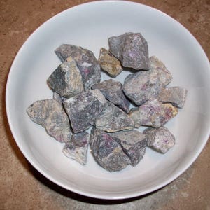 May include: A white bowl filled with a collection of rough, gray and pink stones. The stones have a mottled appearance and are of various sizes.
