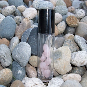 May include: A clear glass roller bottle with a black cap filled with pink rose quartz crystals. The bottle is sitting on a bed of gray and brown rocks.