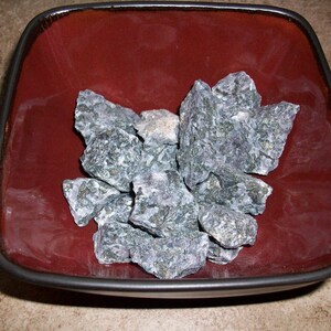 May include: A square, burgundy-colored bowl filled with several rough, gray-green rocks. The bowl has a dark rim and sits on a light-colored surface. The rocks have a textured appearance.
