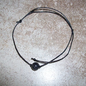 May include: Black beaded necklace with a single, dark, round bead. The necklace is made of thin, black cord, knotted to secure the bead. The necklace is simple and minimalist, suitable for everyday wear.