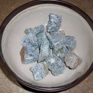 May include: A bowl filled with small, green and gray stones. The stones are smooth and have a natural, rough texture.