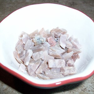 May include: A white bowl with a red rim filled with small, light pink and gray selenite crystal chips.