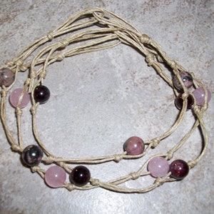 May include: A multi-strand hemp necklace with pink and dark red beads. The necklace is made with natural hemp cord and features a variety of gemstone beads.