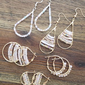 May include: A set of six pairs of gold hoop earrings with delicate beaded accents. The earrings feature a variety of shapes, including teardrop and round hoops. The beads are a mix of iridescent and opaque white.