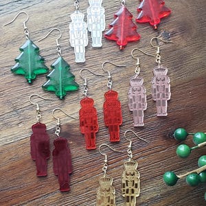 May include: Assortment of Christmas-themed earrings featuring nutcracker and Christmas tree designs. The earrings are made of acrylic and come in a variety of colors, including red, green, gold, silver, and pink.