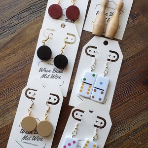 May include: Five pairs of earrings on white cards with the text "When Bead Met Wire" and "Handmade Goods". The earrings are made of wood, plastic, and metal. The designs include red and black circles, bowling pins, dominoes, and a tan circle.