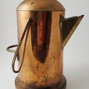 May include: A vintage copper coffee pot with a rounded lid and a spout. The pot has a handle and a small knob on top. The copper has a warm, aged patina, suggesting it's an antique or a well-loved item.