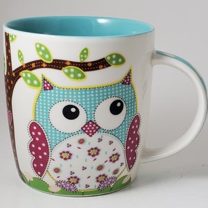 May include: White ceramic mug with a teal interior and a large owl design. The owl is primarily blue with polka dots, and has pink, yellow, and green accents. A brown tree branch with green leaves is also featured.