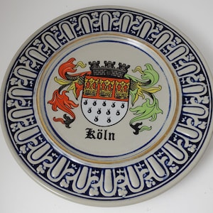 May include: A ceramic plate with a blue and white border and a gold rim. The center of the plate features a colorful coat of arms with a castle, crowns, and flames. The text "Köln" is written below the coat of arms.