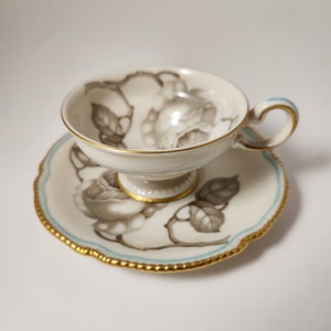 May include: A white porcelain teacup and saucer set with a delicate floral design. The cup and saucer feature a gray and white rose pattern with gold trim.