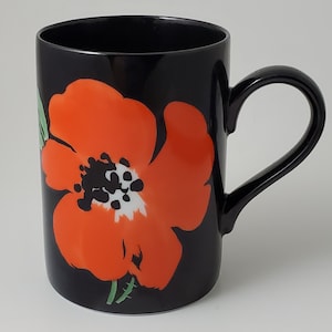 May include: Black ceramic mug with a large orange poppy flower design. The mug has a white center with black dots and a green stem.