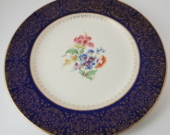 May include: A round, white ceramic plate with a navy blue border featuring a gold filigree design. The center of the plate displays a bouquet of colorful flowers, including pink, blue, and orange blooms. The plate has a gold rim.
