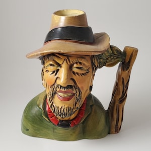 May include: A ceramic mug shaped like a smiling man wearing a hat and a red necklace. The mug has a handle that looks like a wooden branch. The man's face is detailed with a beard and wrinkles, and the hat has a black band.