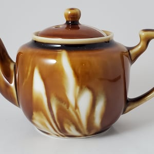 Puede incluir: Una tetera de cerámica marrón con un diseño de remolino blanco. La tetera tiene una tapa con un pequeño agujero en la parte superior.